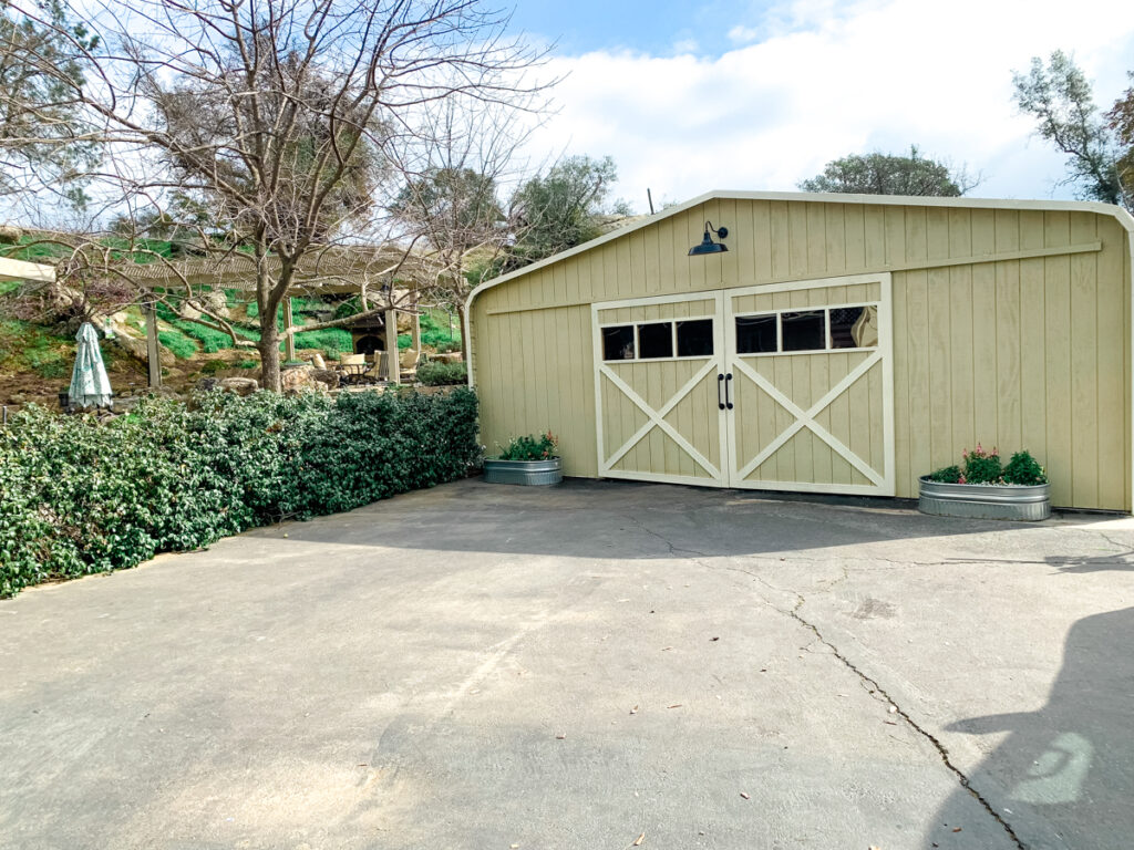 Completed carport barn photo. Large barn light to illuminate the barn doors. The barn doors have windows in them and an X pattern on the front with large metal handles. Two metal troughs are placed in front of barn with seasonal flowers. 
