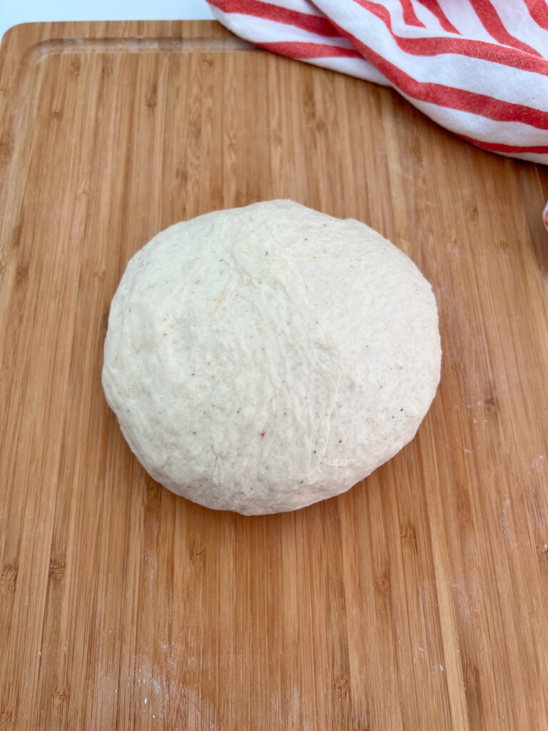 The photo shows a dough ball ready to be shaped into a pizza crust on top of a cutting board.